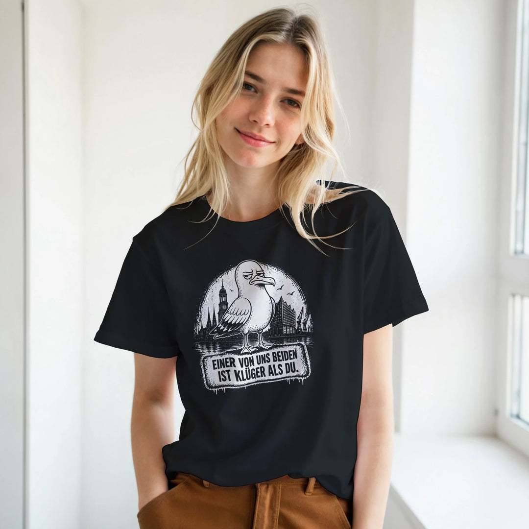 Blonde Frau trägt das schwarze Shirt mit Möwen-Print vor heller Fensterwand, kombiniert mit brauner Hose.