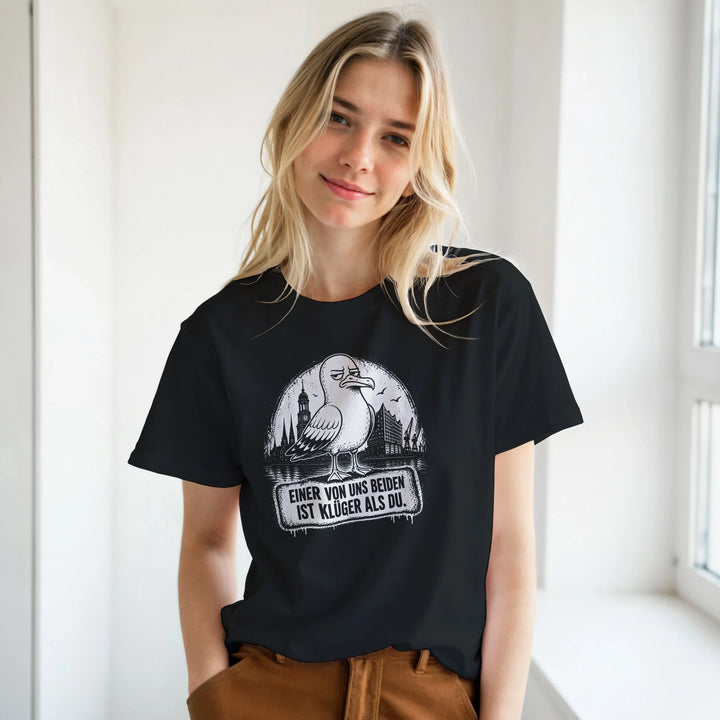Blonde Frau trägt das schwarze Shirt mit Möwen-Print vor heller Fensterwand, kombiniert mit brauner Hose.