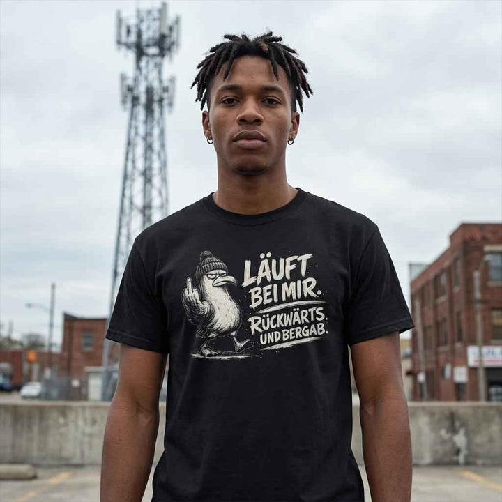 Mann trägt schwarzes T-Shirt mit Möwen-Print „Läuft bei mir. Rückwärts. Und bergab.“ auf einem Parkdeck vor Sendemast.