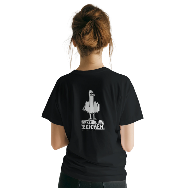 Rückansicht einer Frau mit Dutt, Möwen-Stinkefinger-Print und Schriftzug „Erkenne die Zeichen“ auf dem Rücken des schwarzen T-Shirts.