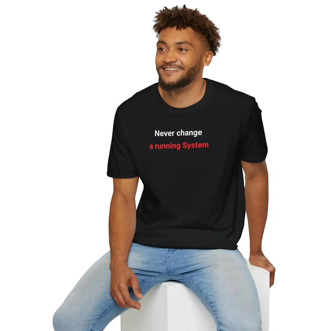 Lächelnder Mann sitzt auf einem weißen Würfel und blickt zur Seite, trägt das schwarze „Never change a running System“-Shirt, Studiofoto.