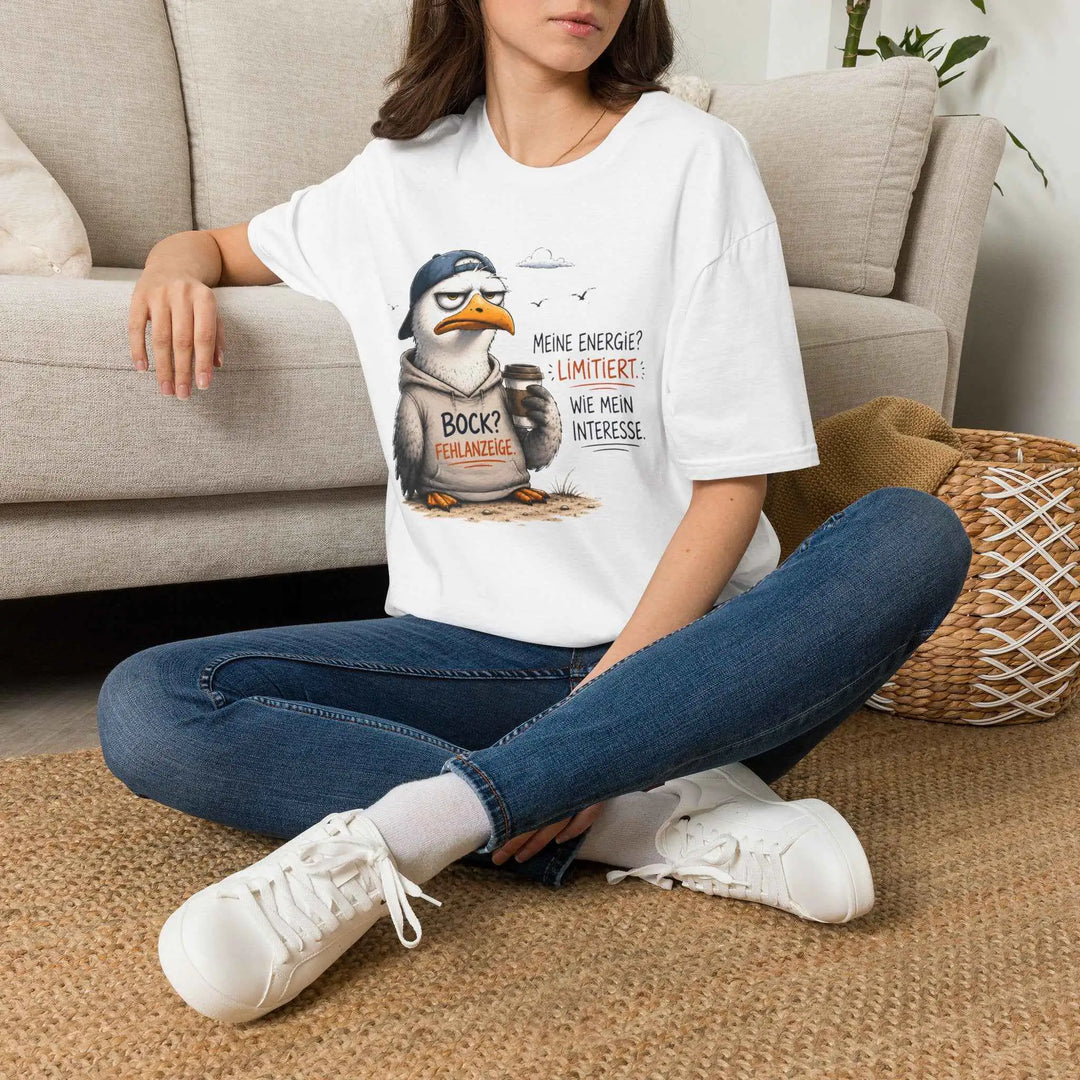 Frau sitzt locker auf dem Boden vor einem Sofa und trägt weißes T-Shirt mit Illustration einer genervten Möwe und dem Spruch „Bock? Fehlanzeige.“, Jeans und Sneakers.