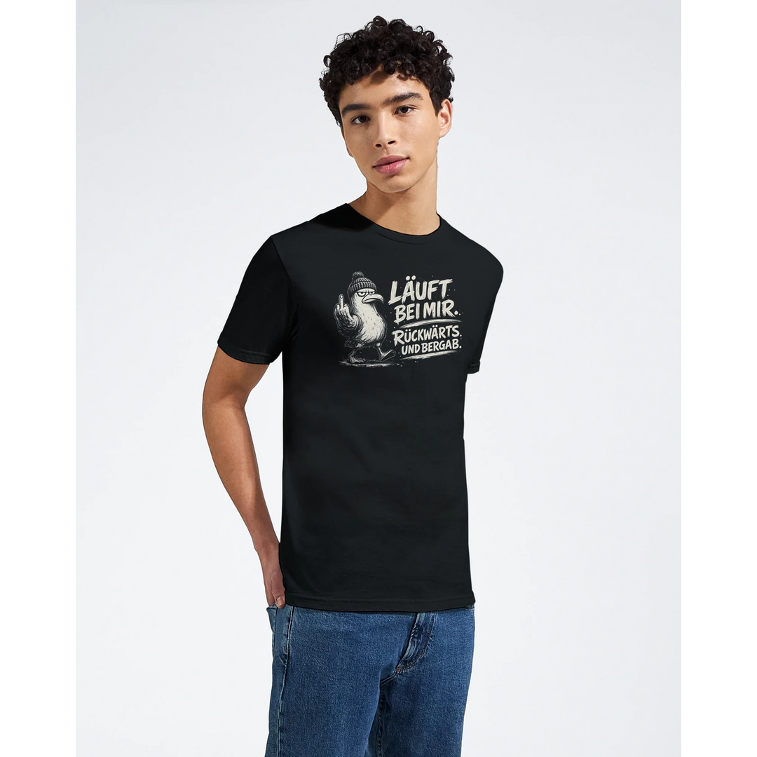 Model im schwarzen Möwen-Shirt mit Spruch „Läuft bei mir. Rückwärts. Und bergab.“ vor weißem Studiohintergrund.