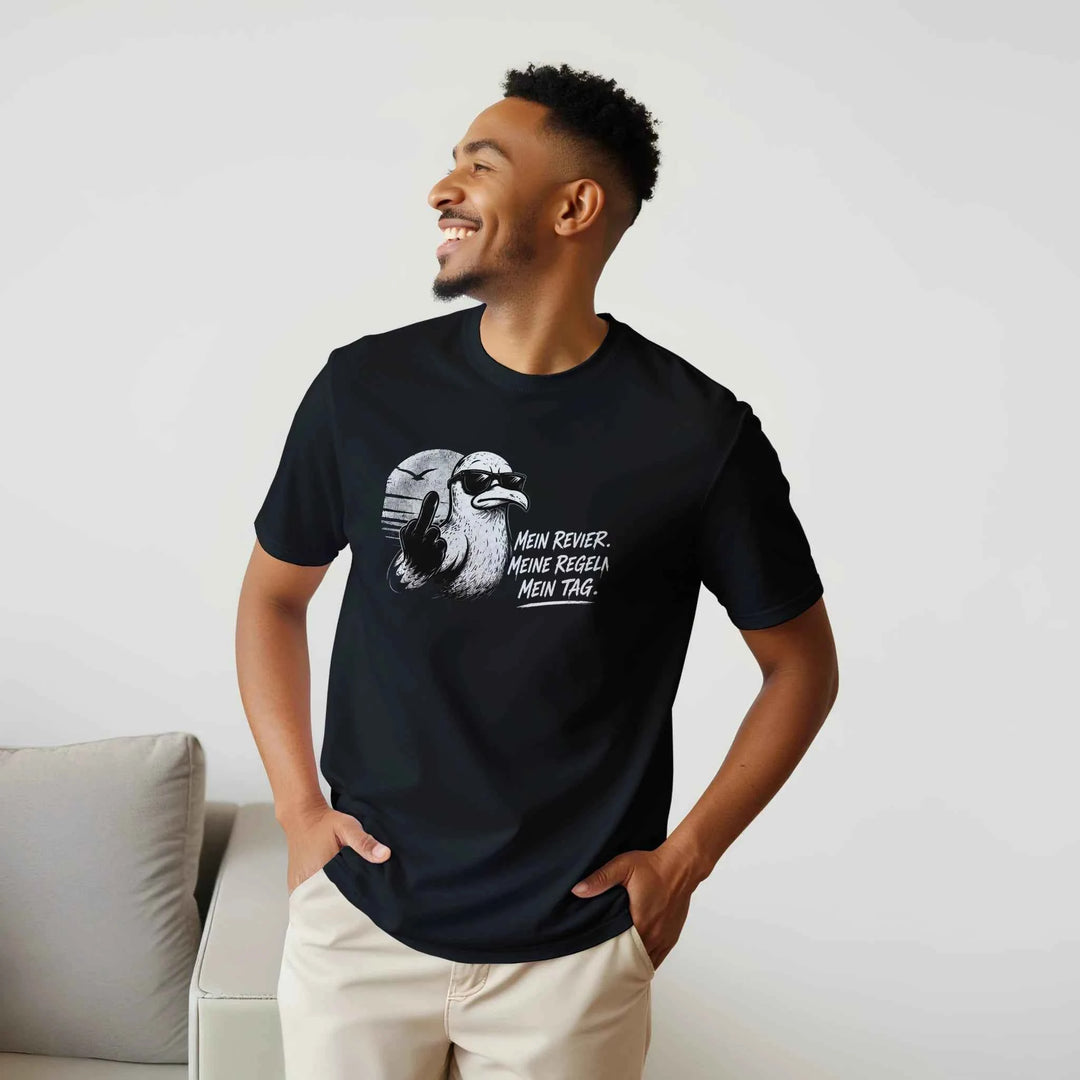 Mann in schwarzem T-Shirt mit Möwen-Motiv „Mein Revier. Meine Regeln. Mein Tag.“ lehnt lächelnd an einem Sofa