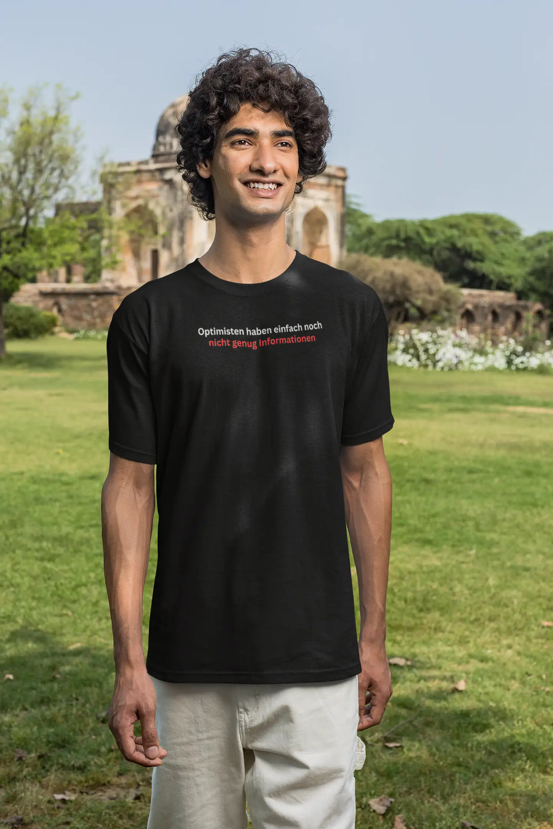 Lächelnder Mann auf Wiese vor historischem Gebäude im schwarzen „Optimisten“-T-Shirt