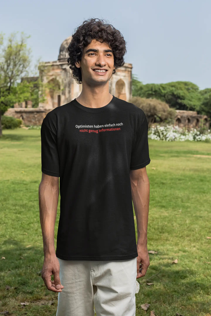 Lächelnder Mann auf Wiese vor historischem Gebäude im schwarzen „Optimisten“-T-Shirt