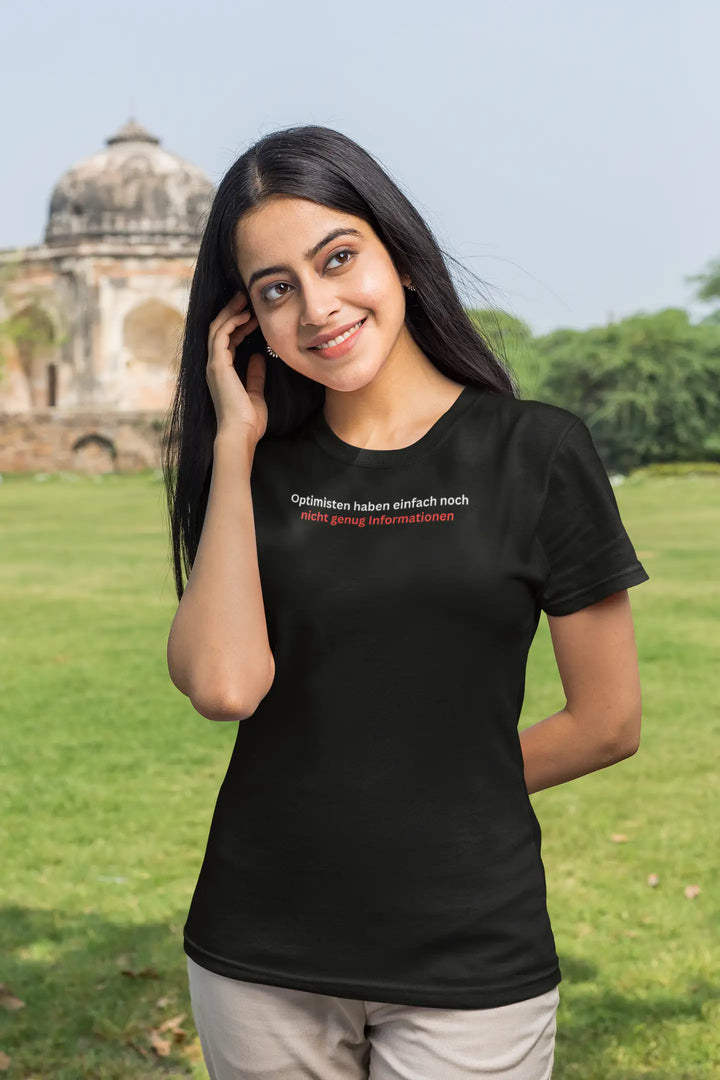 Frau lächelt auf Wiese vor historischem Gebäude im schwarzen „Optimisten“-T-Shirt