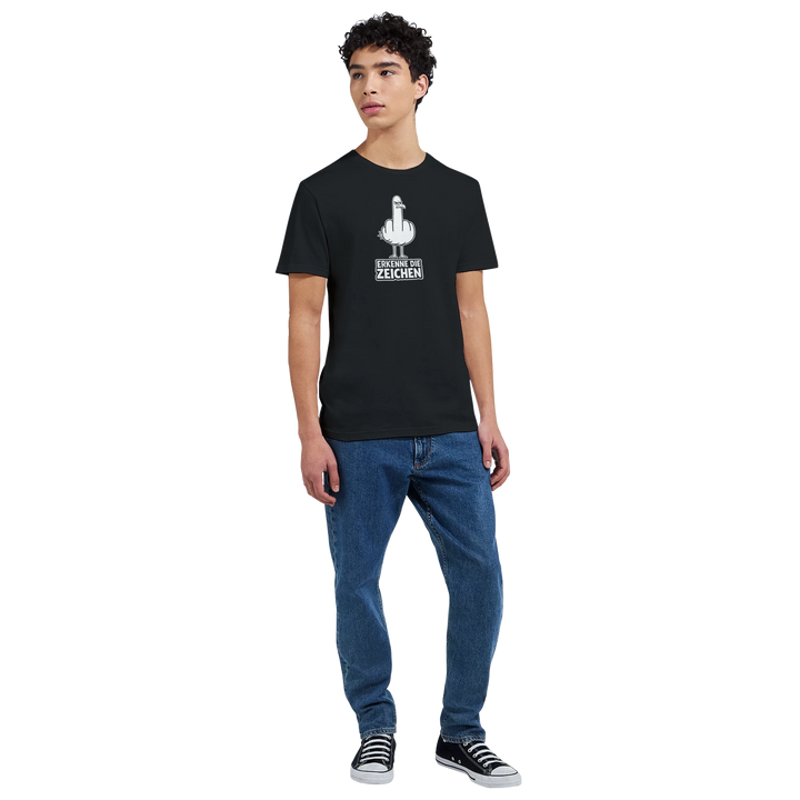 Ganzkörperaufnahme eines Mannes in Jeans und Sneakers, schwarzes T-Shirt mit Möwen-Print „Erkenne die Zeichen“ auf der Brust.