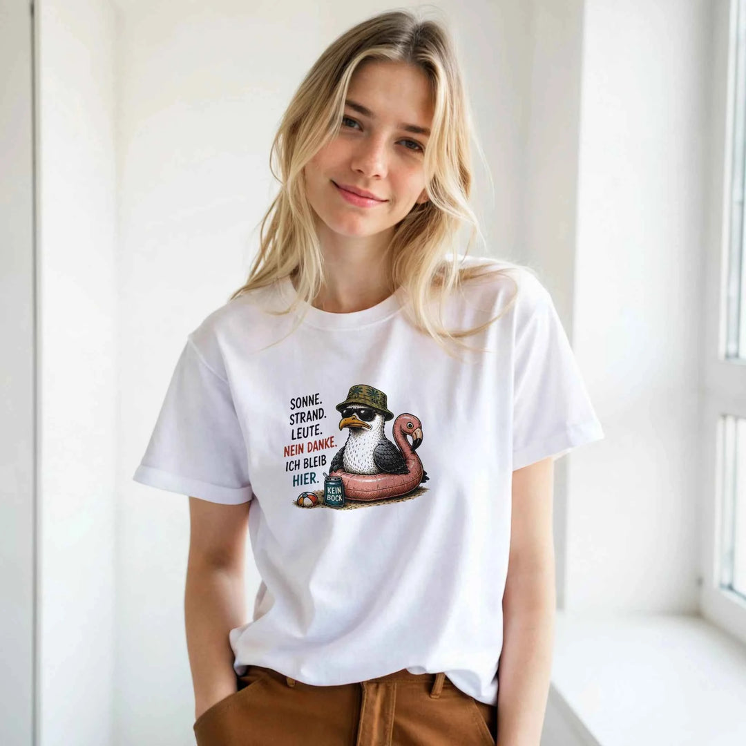 Blonde Frau trägt das weiße T-Shirt mit Möwen-Print vor heller Fensterwand, kombiniert mit brauner Hose.