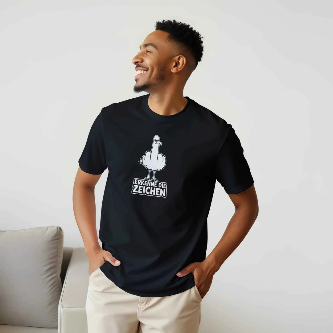 Lächelnder Mann im Halbprofil zeigt das schwarze T-Shirt mit Möwen-Stinkefinger-Motiv und Schriftzug „Erkenne die Zeichen“.