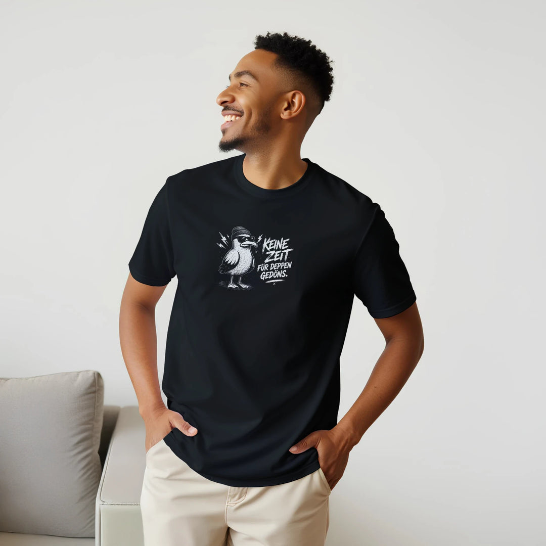 Lächelnder Mann steht im Wohnzimmer neben einem Sofa, schwarzes T-Shirt mit Möwen-Print „Keine Zeit für Deppen-Gedöns.“