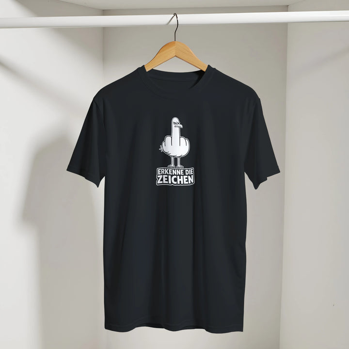 Schwarzes T-Shirt mit Möwen-Print hängt am Kleiderbügel in der Zimmerecke, Spruch „Erkenne die Zeichen“.