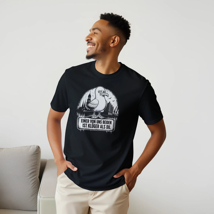 Lächelnder Mann im schwarzen T-Shirt mit Hamburg-Möwe entspannt vor einem grauen Sofa.