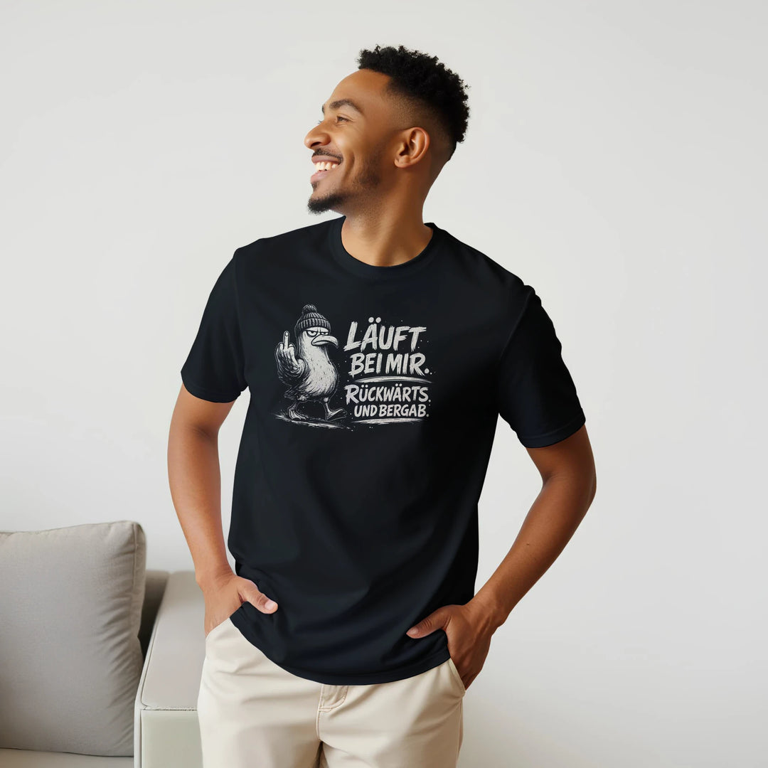 Lächelnder Mann trägt das schwarze Möwen-Shirt mit den Händen in den Taschen vor einem Sofa.