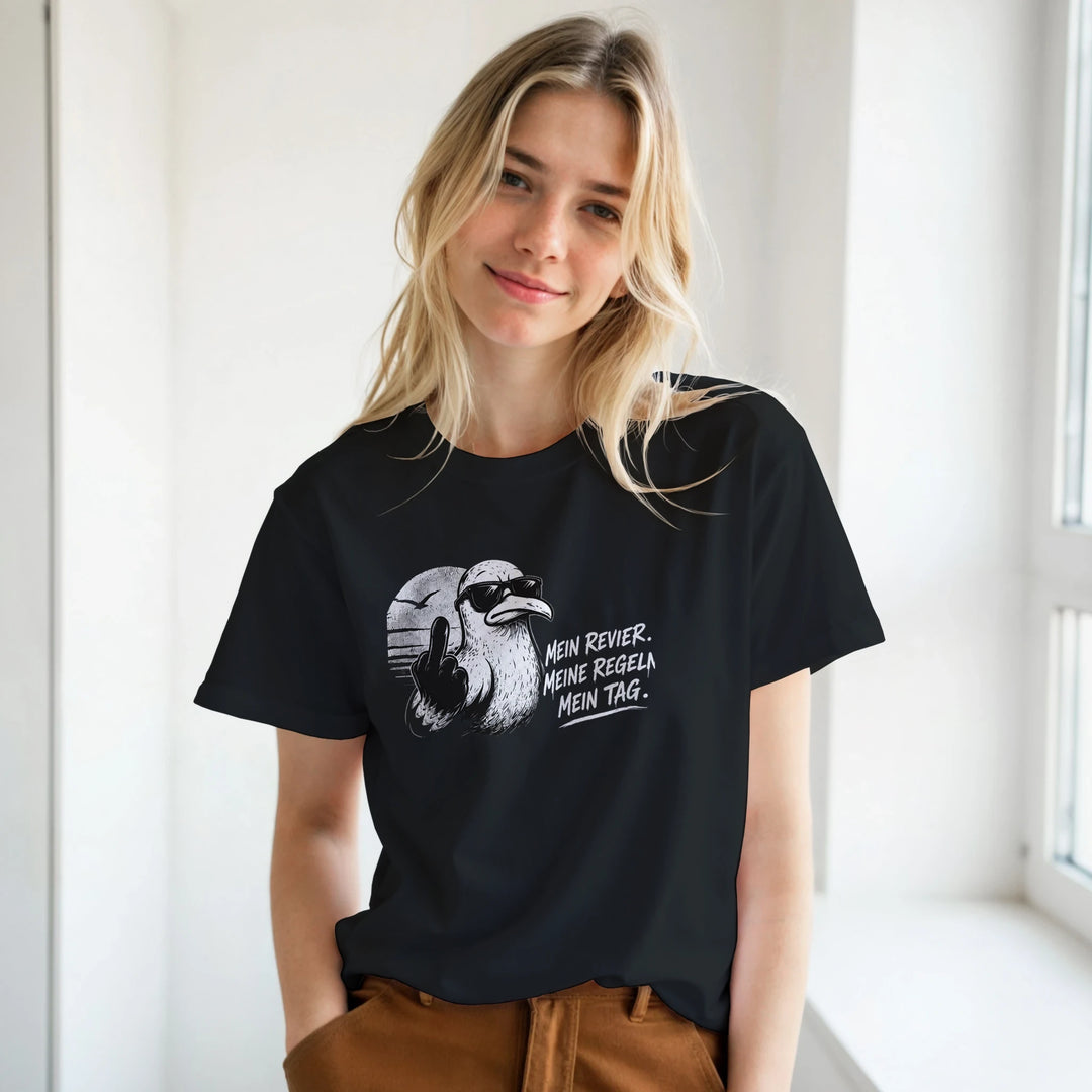 Blonde Frau in schwarzem T-Shirt mit frechem Möwen-Print, lächelnd am Fenster stehend