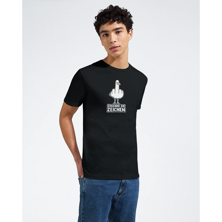 Junger Mann posiert von vorne im Studio, schwarzes T-Shirt mit weißem Möwen-Print und Spruch „Erkenne die Zeichen“.