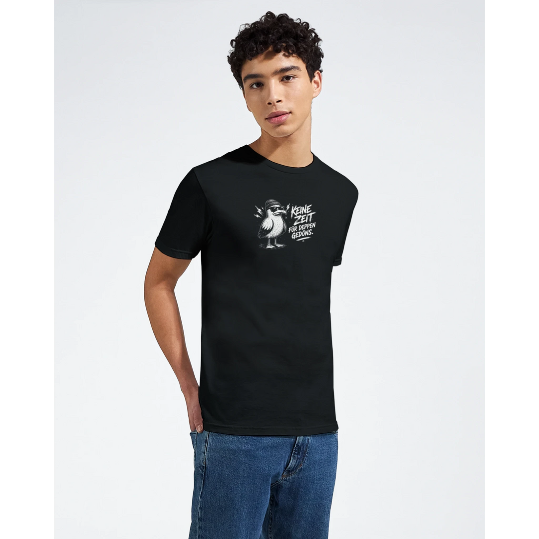 Junger Mann im Halbprofil trägt schwarzes T-Shirt mit Möwen-Print „Keine Zeit für Deppen-Gedöns.“ vor grauem Studiohintergrund.