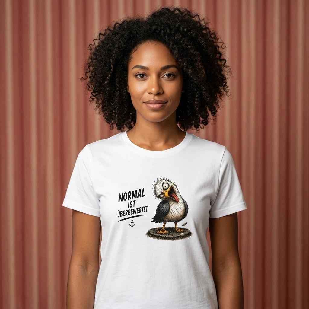 Frau mit lockigem Haar in weißem T-Shirt mit schräger Möwe und Spruch „Normal ist überbewertet.“ vor gestreiftem Hintergrund