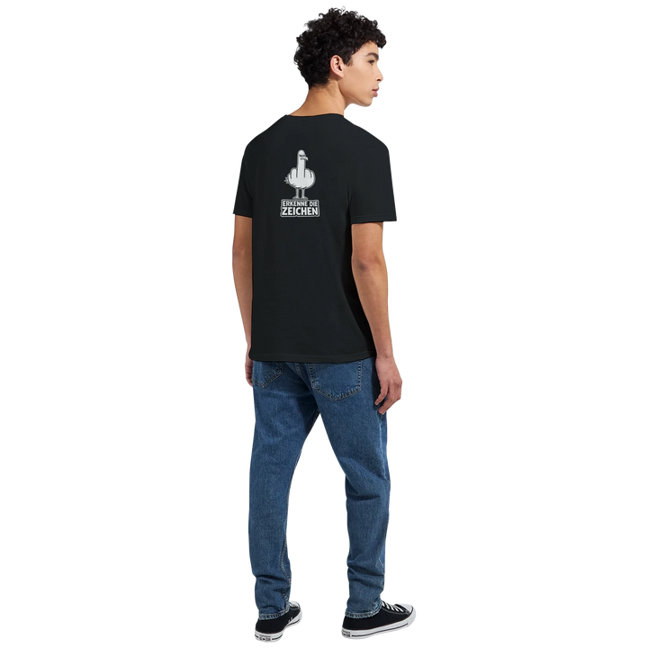 Rückansicht eines Mannes beim Gehen auf der Straße, Möwen-Print und Spruch „Erkenne die Zeichen“ auf dem Rücken des schwarzen T-Shirts.