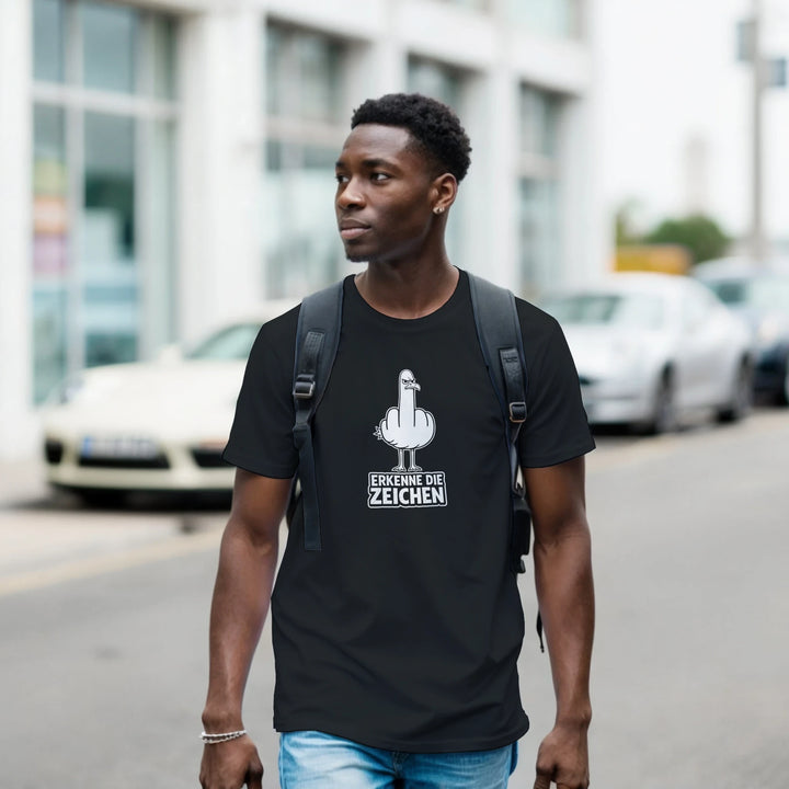 Mann mit Rucksack geht durch die Stadt, schwarzes T-Shirt mit Möwen-Stinkefinger-Motiv und Spruch „Erkenne die Zeichen“.