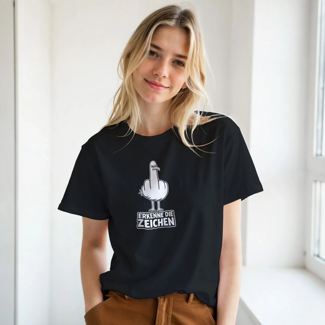 Blonde Frau lächelt am Fenster, schwarzes T-Shirt mit Möwen-Print und Schriftzug „Erkenne die Zeichen“.