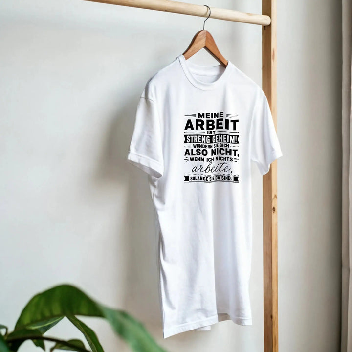 Weißes „Streng geheim“ T-Shirt hängt am Kleiderbügel neben Pflanze