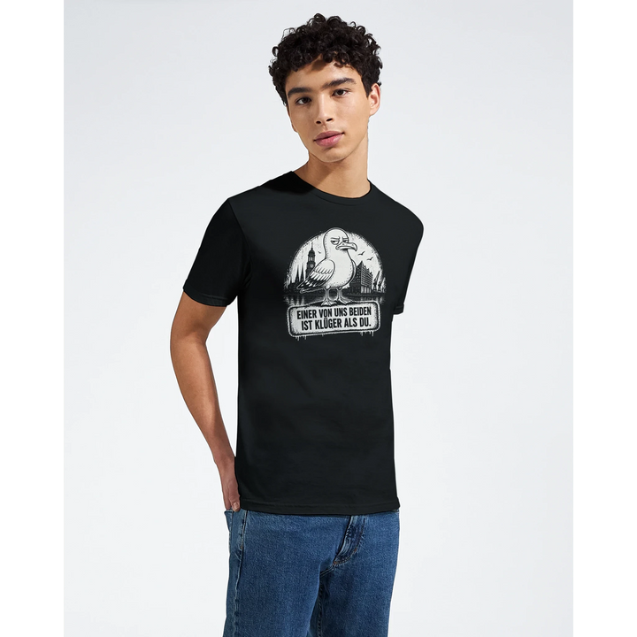 Mann mit lockigem Haar trägt das schwarze T-Shirt „Einer von uns beiden ist klüger als du“ vor weißem Studiohintergrund.