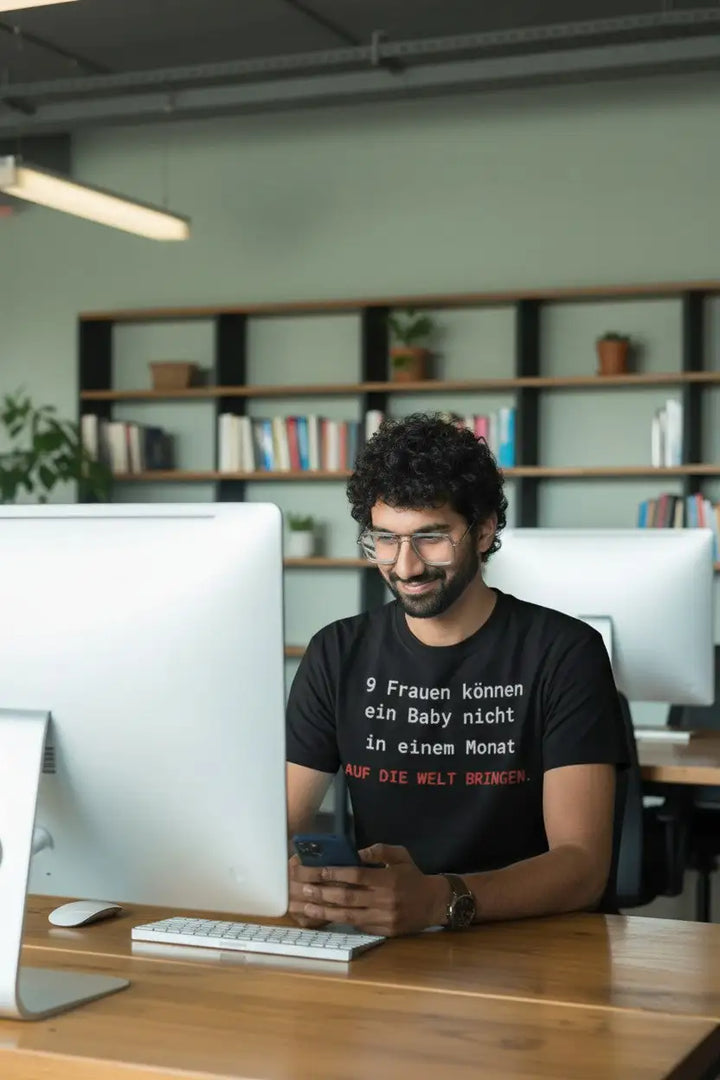 Mann sitzt lächelnd am Schreibtisch mit zwei iMacs und Smartphone in der Hand, trägt schwarzes T-Shirt mit Brooks-Law-Spruch