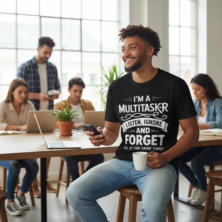 Lächelnder Mann sitzt mit Kaffeetasse und Smartphone in einem Café-Büro, im Hintergrund Kollegen an Laptops, trägt schwarzes T-Shirt mit Aufdruck „I&