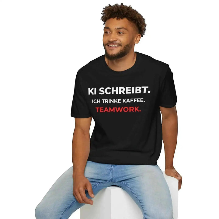 Lächelnder Mann sitzt auf einem Hocker und trägt schwarzes T-Shirt mit Aufdruck „KI schreibt. Ich trinke Kaffee. Teamwork.“