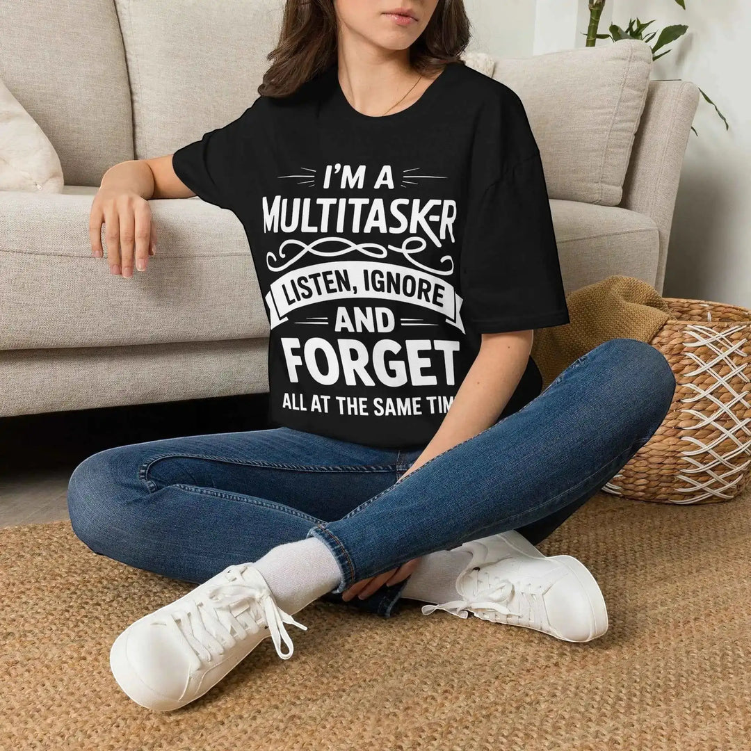 Frau sitzt locker auf dem Boden vor einem Sofa und trägt das schwarze Multitasker-T-Shirt mit Jeans und Sneakers.